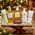 A Christmas-themed gift bath and body set arranged in a gold tray on top of a wrapped gift box. The set includes White Compass Meridian Shave Oil, Afterglow Deep Cleansing Oil, Magnesium & Lavender Lotion, Ember Botanicals Theramint and Sunshine Lotions, and a bar of Spiced Cider soap.